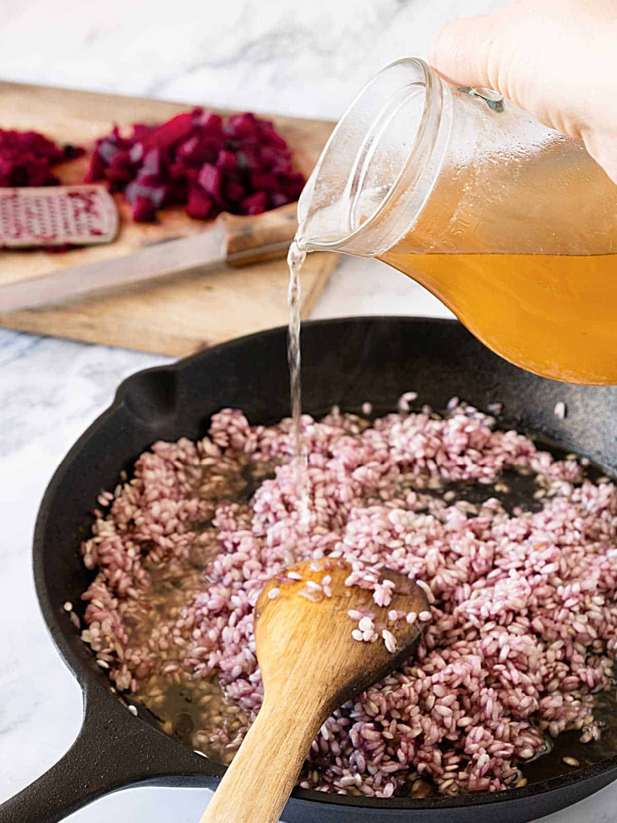 Adding broth from a jug to beet risotto in a cast-iron skillet. Wooden spoon inside.
