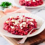 Wooden board with white plate of goat cheese beet risotto. Pink cloth, white marble surface, chopped chives.