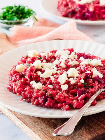 Wooden board with white plate of goat cheese beet risotto. Pink cloth, white marble surface, chopped chives.