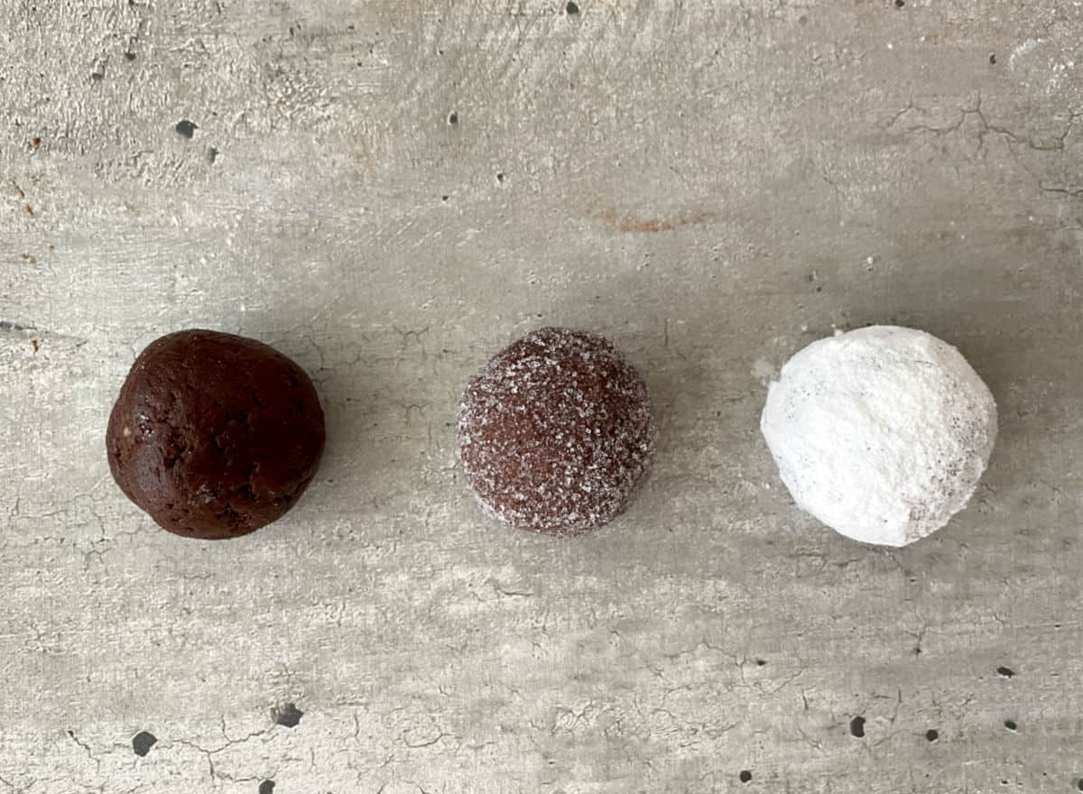 Grey surface with three chocolate cookie dough balls: plain, sugar coated, and powdered sugar coated. 
