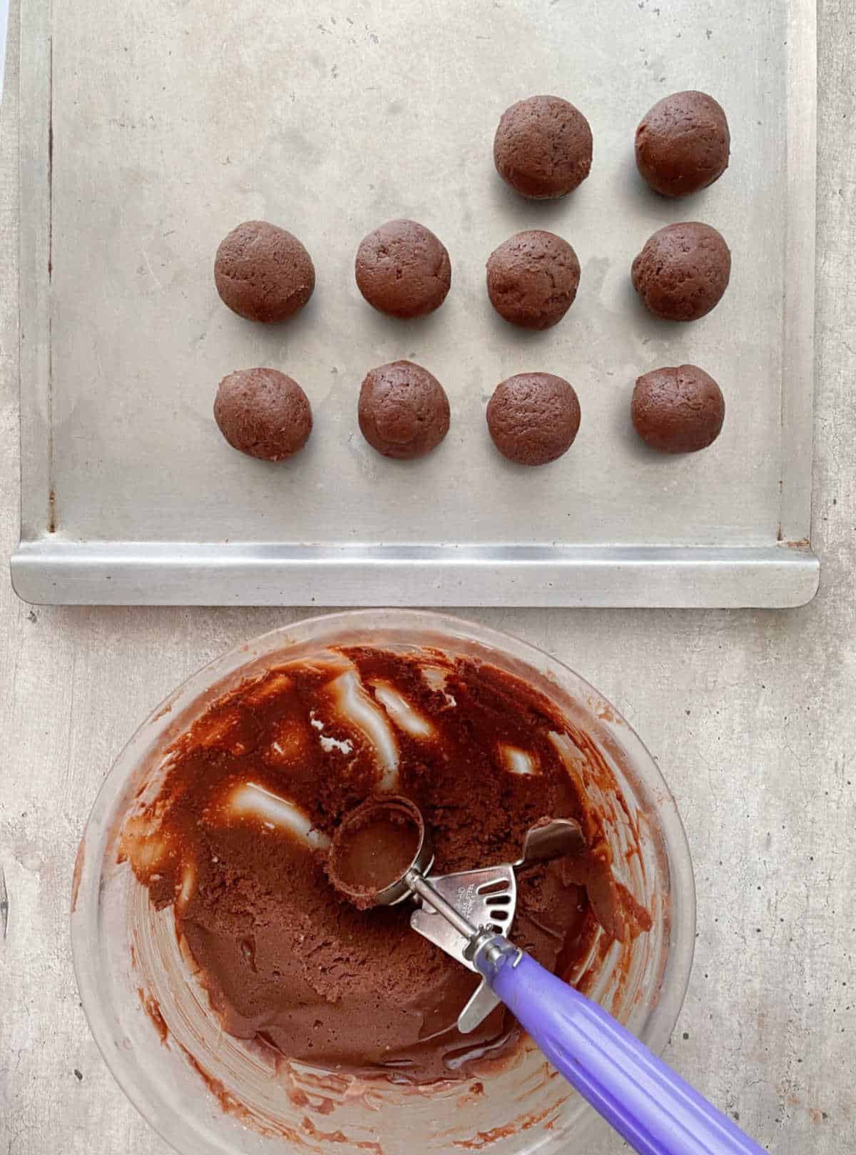 Baking sheet with chocolate cookie dough rounds. Glass bowl with dough and scoop. Grey surface.