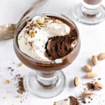White surface with cream-topped French chocolate mousse. Bite missing. Silver spoon, loose pistachios.