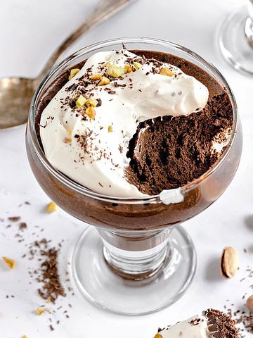 White surface with cream-topped French chocolate mousse. Bite missing. Silver spoon, loose pistachios.