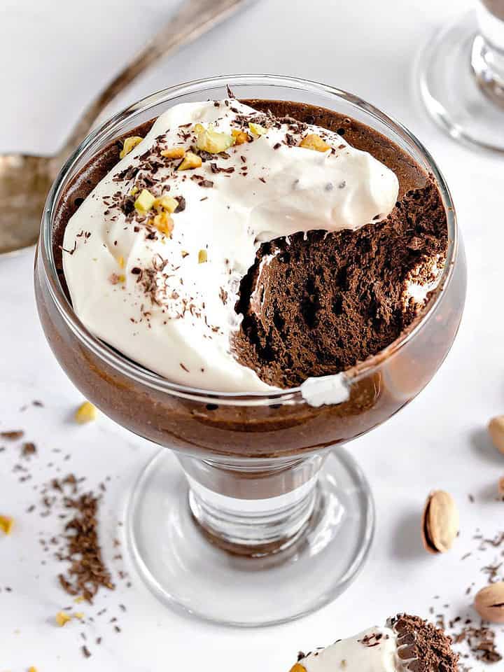 White surface with cream-topped French chocolate mousse. Bite missing. Silver spoon, loose pistachios.