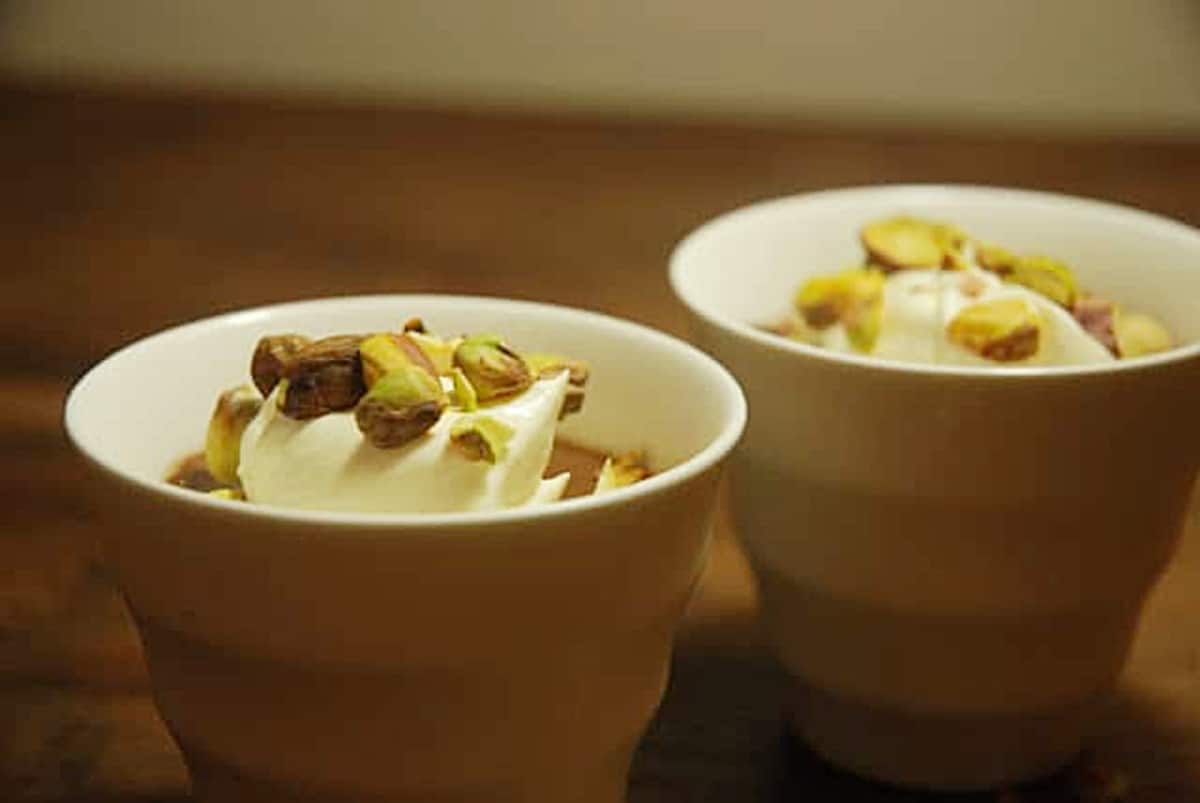 Two white cups showing whipped cream and pistachios. Wooden background. 