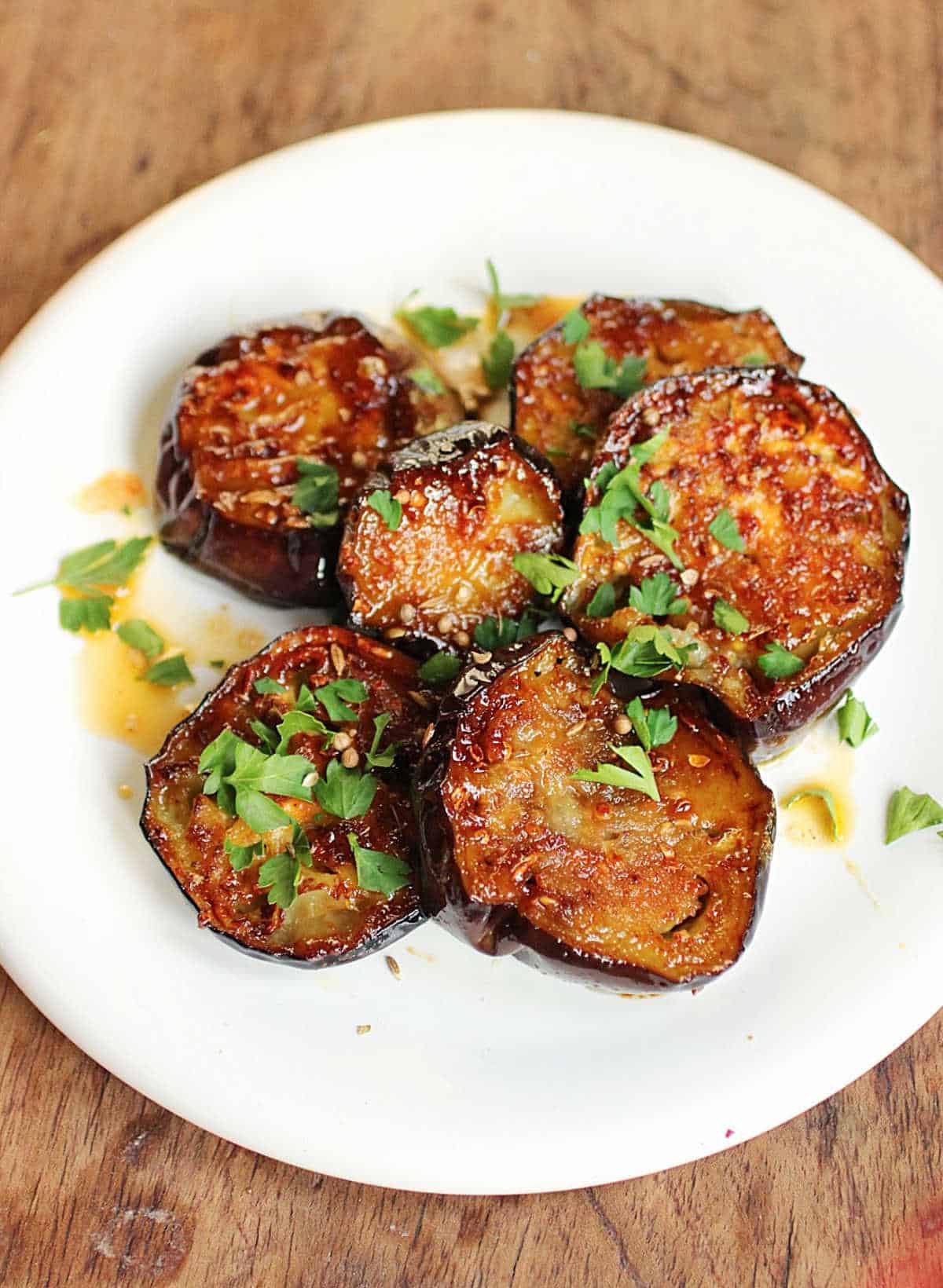 Eggplant rounds with caramelized coating and chopped herbs on a white plate on a wooden table.