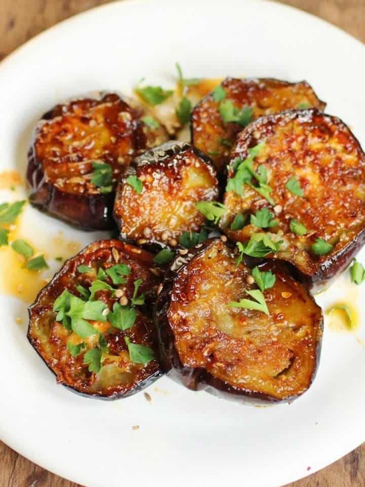 Several eggplant rounds with honey and chopped parsley on a white plate set on a wooden surface.