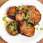 A white plate with honey eggplant slices garnished with chopped herbs on a wooden table.