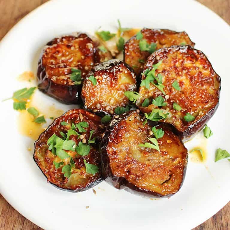 A white plate with honey eggplant slices garnished with chopped herbs on a wooden table.