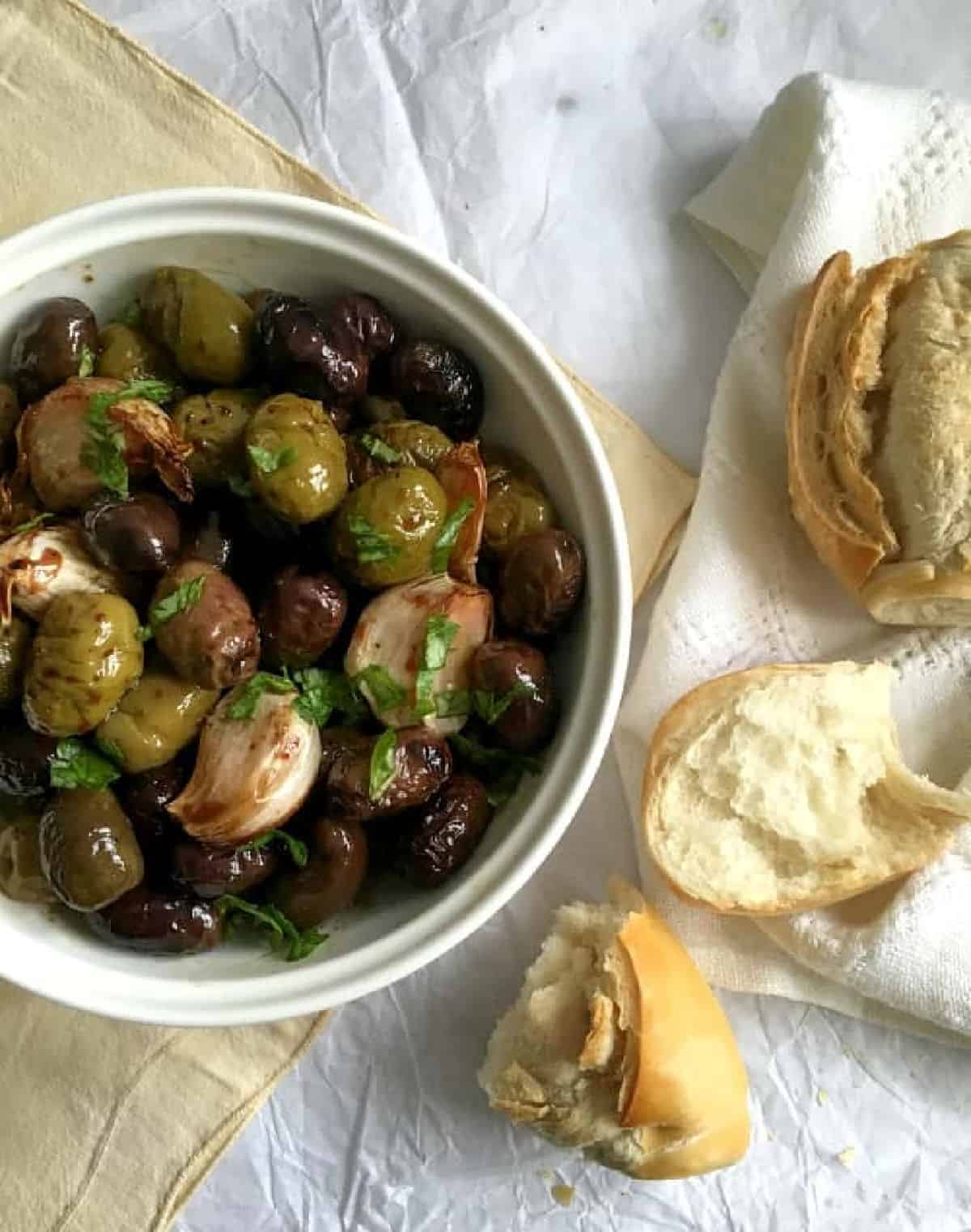 Pieces of french bread beside white bowl with olives and garlic on a white and beige cloth. 