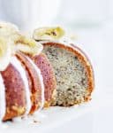 Slice of banana cake pulled out from whole glazed bundt, banana chips on top, white background