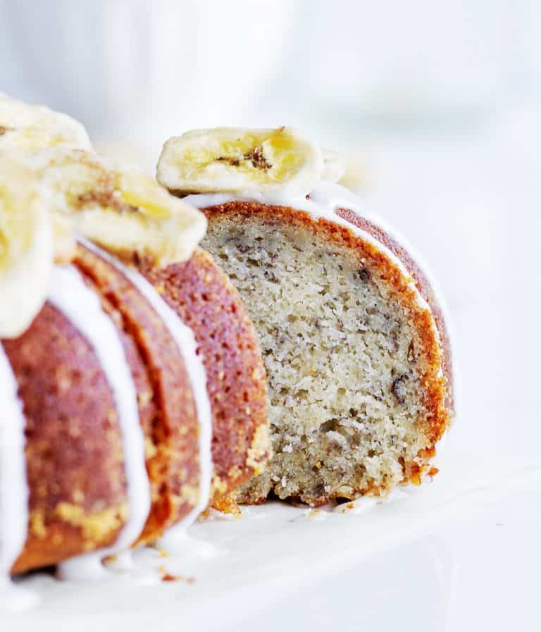 Slice of banana cake pulled out from whole glazed bundt, banana chips on top, white background