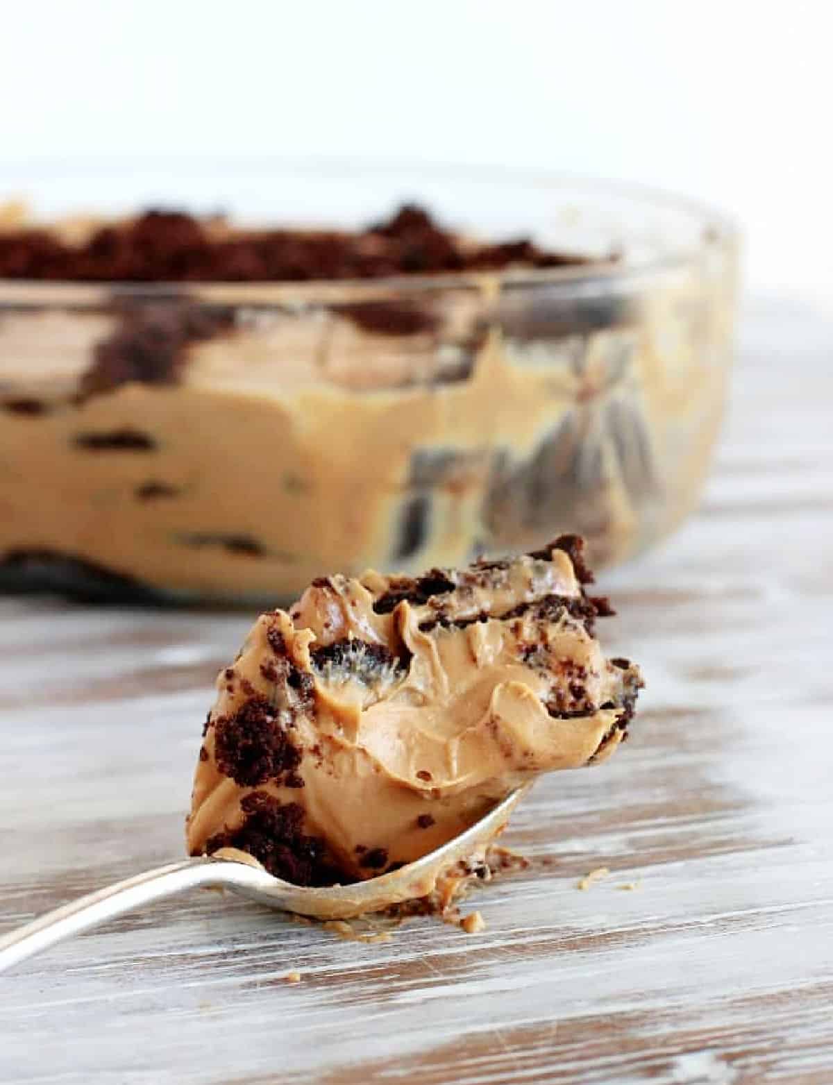 Spoon with chocotorta on whitish wooden table. Glass bowl with dessert in background.