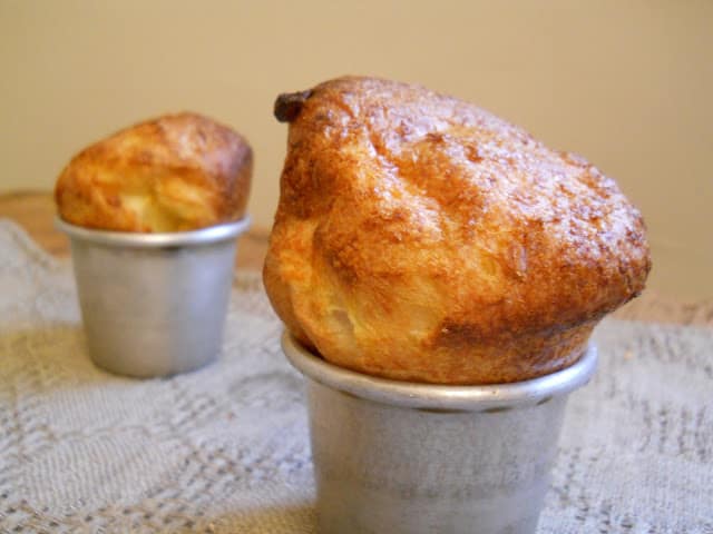Two popovers in metal pan on grey cloth. Brownish yellow wall as background. 