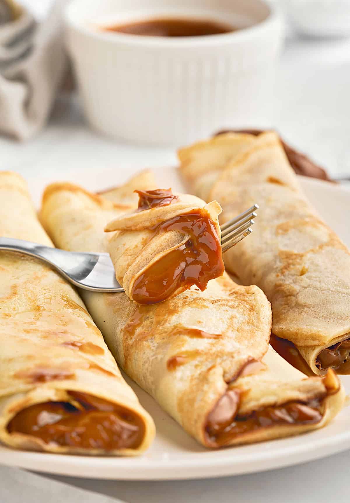 Forked bite of dulce de leche crepe on more rolled pancakes. White plate and background.