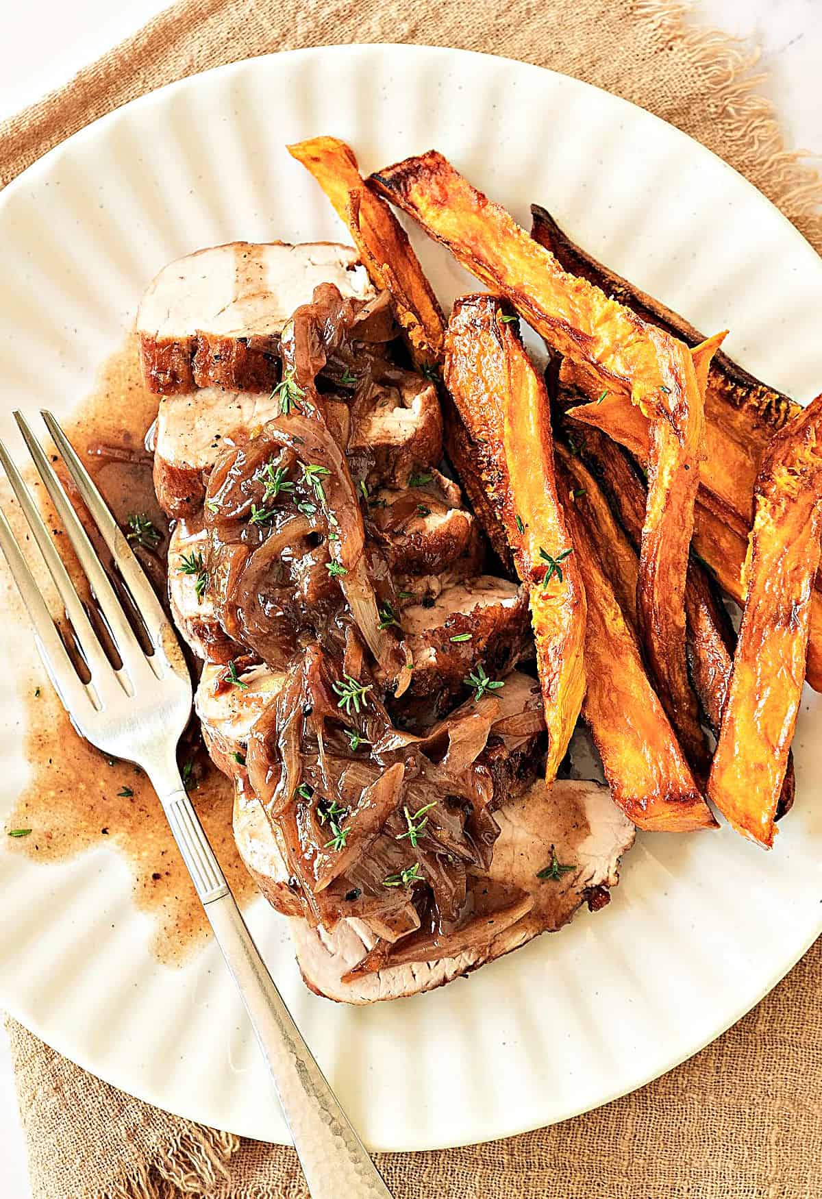 Top view of white plate with sweet potato fries and sliced pork loin with shallots. Silver fork. Beige placeholder.