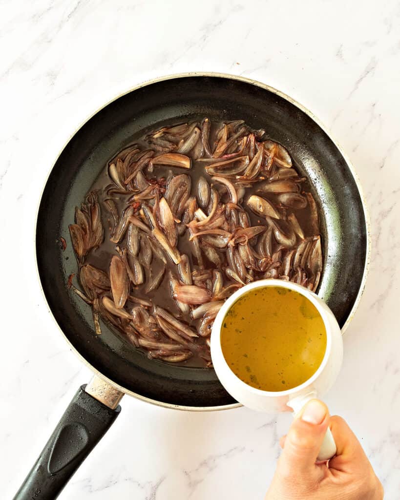 Adding broth to shallot sauce in a black skillet. Top view. White background.