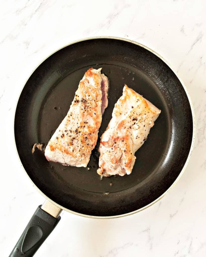 Seared pork tenderloin pieces in a black skillet. White background, top view.