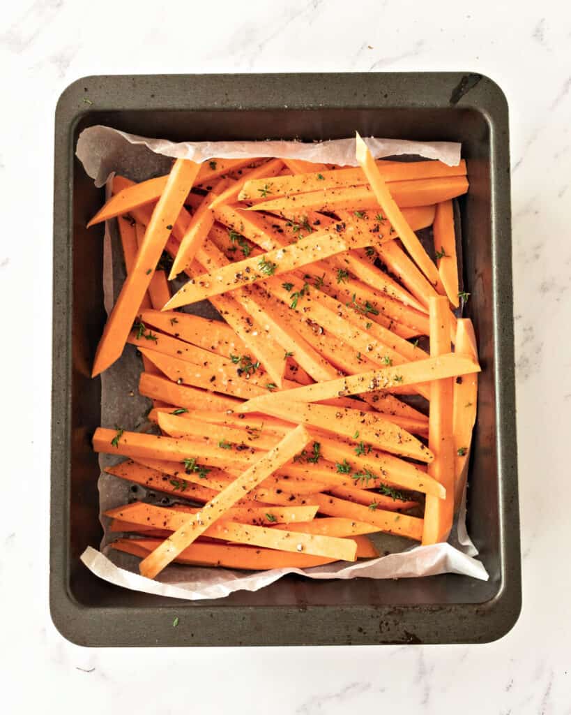 Seasoned sweet potato sticks on parchment paper on black oven tray. Top view. White background. 