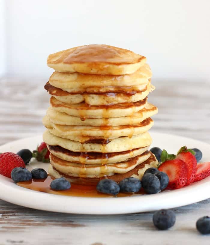 Tall stack of round pancakes on a white plate with berries, syrup dripping, white background