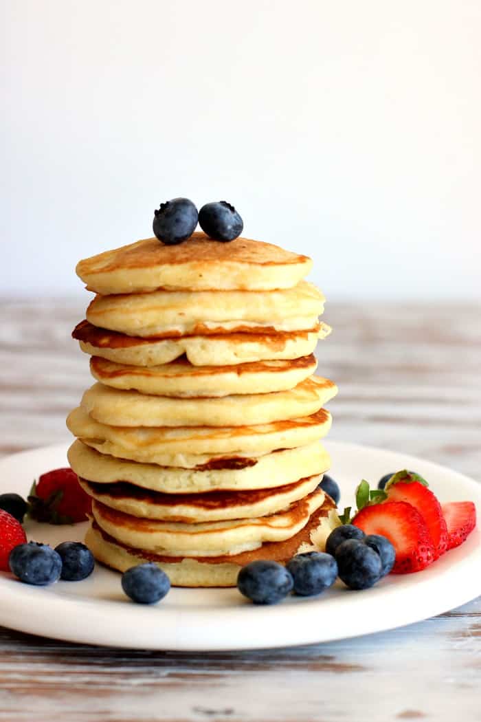 Berries top and around a stack of pancakes on a white plate, white table.