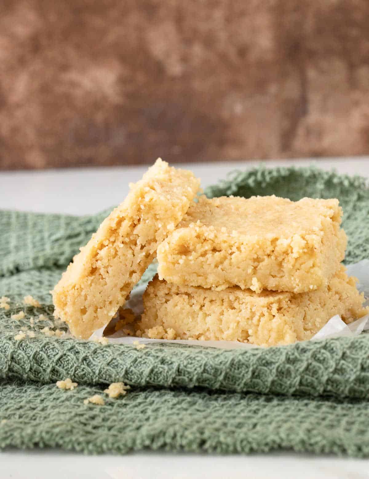 Shortbread squares leaning and stacked on a green cloth with a brown background.