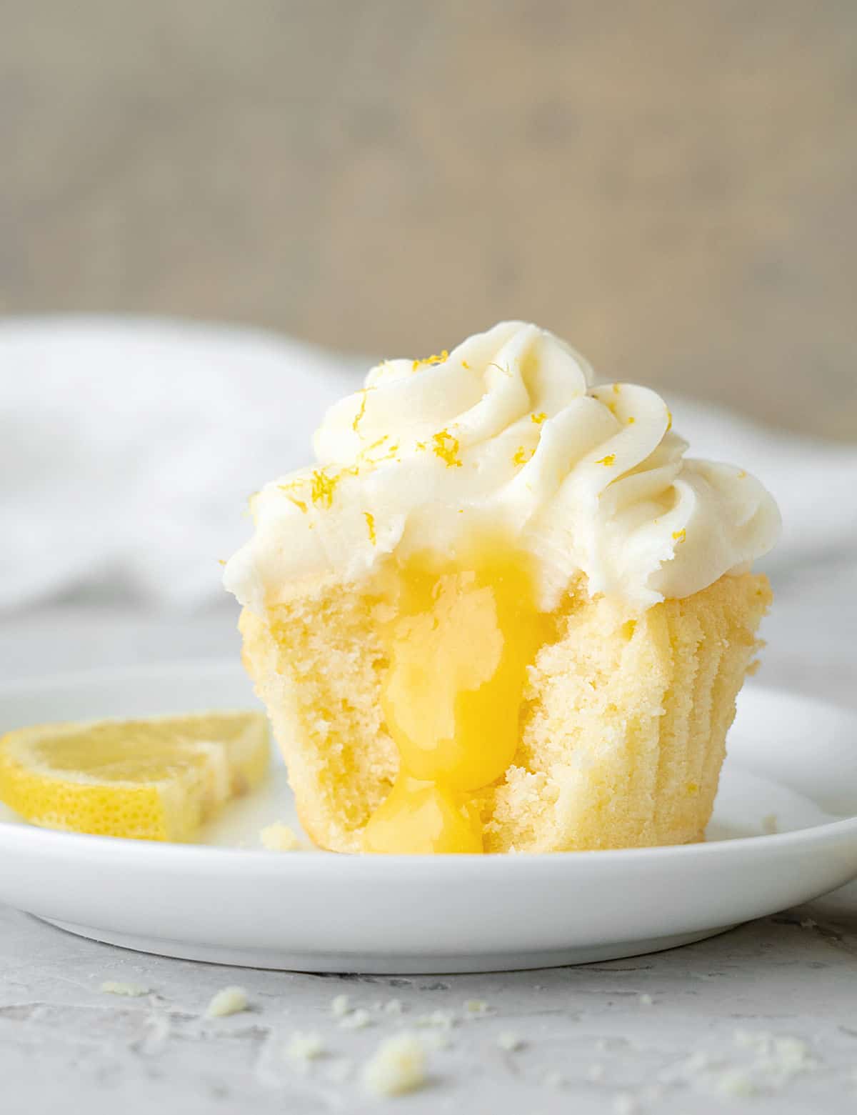 Half frosted lemon curd filled cupcake on a white plate. Beige and white background and grey surface. 