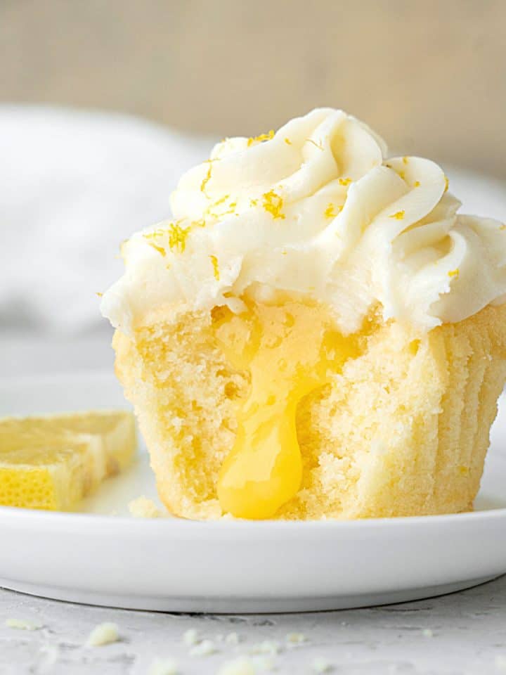 Frosted lemon curd cupcake half on a white plate. White and beige background, grey surface.