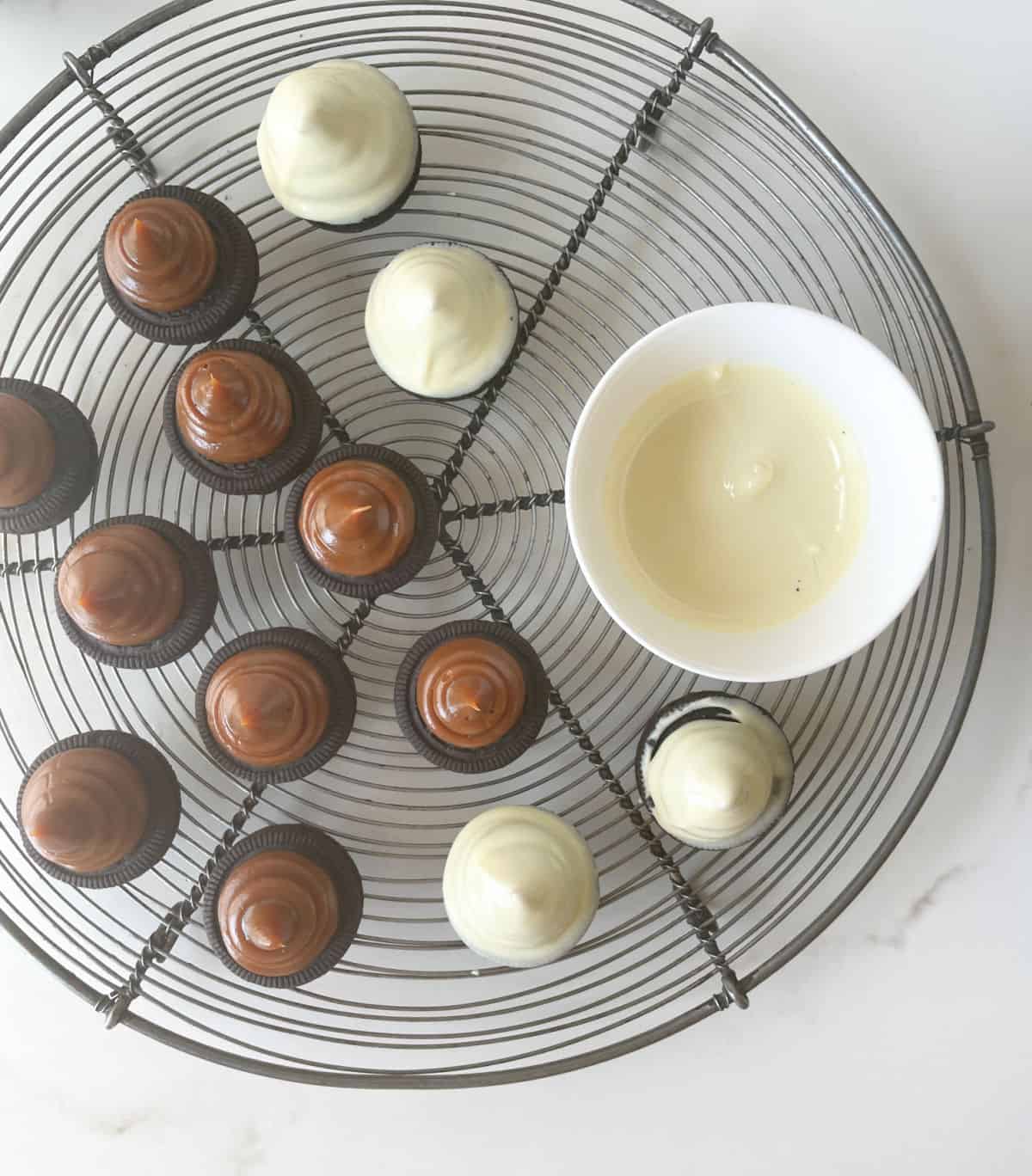 Top view of wire rack with dulce de leche topped oreo cookies, some covered in white chocolate. White bowl with melted chocolate.