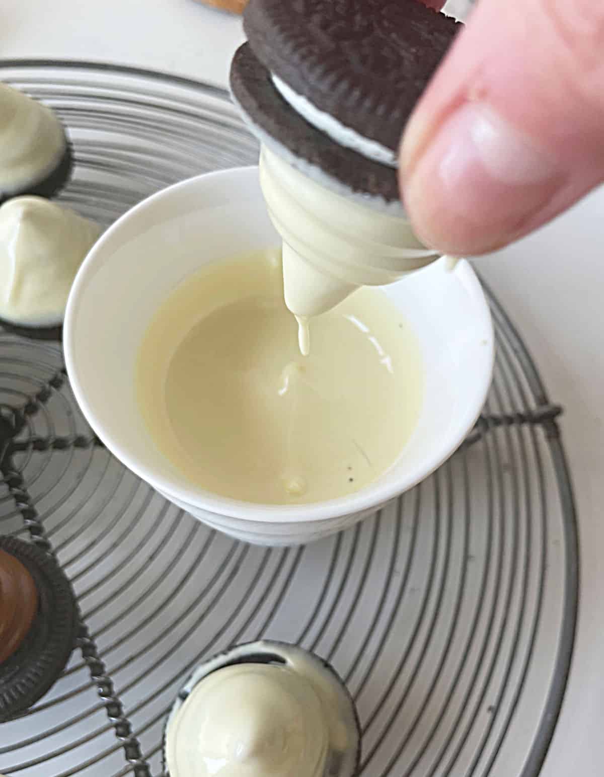 Dipping an oreo cookie with dulce de leche in melted white chocolate in a white bowl on a wire rack.