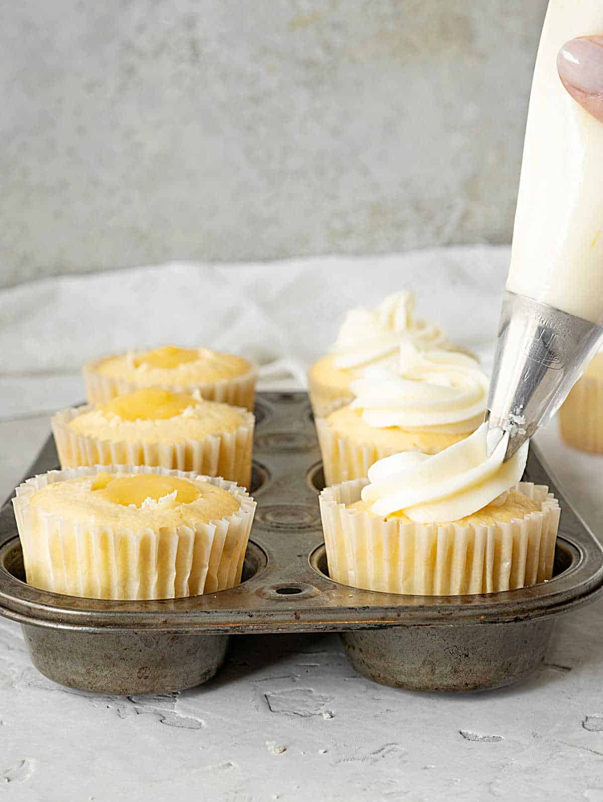 Piping lemon frosting on top of curd filled cupcakes in a metal tin. Grey surface and background.