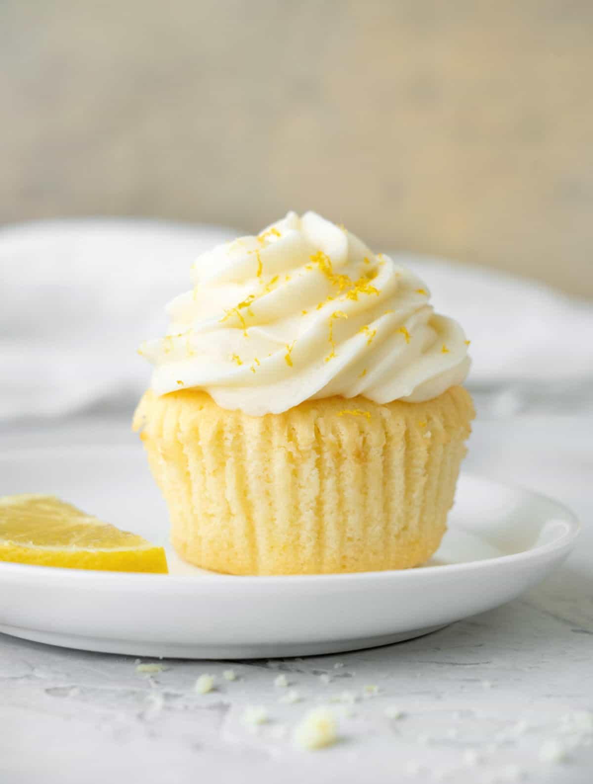 Single frosted lemon cupcake on a white plate. Grey surface and beige background with a white cloth. 