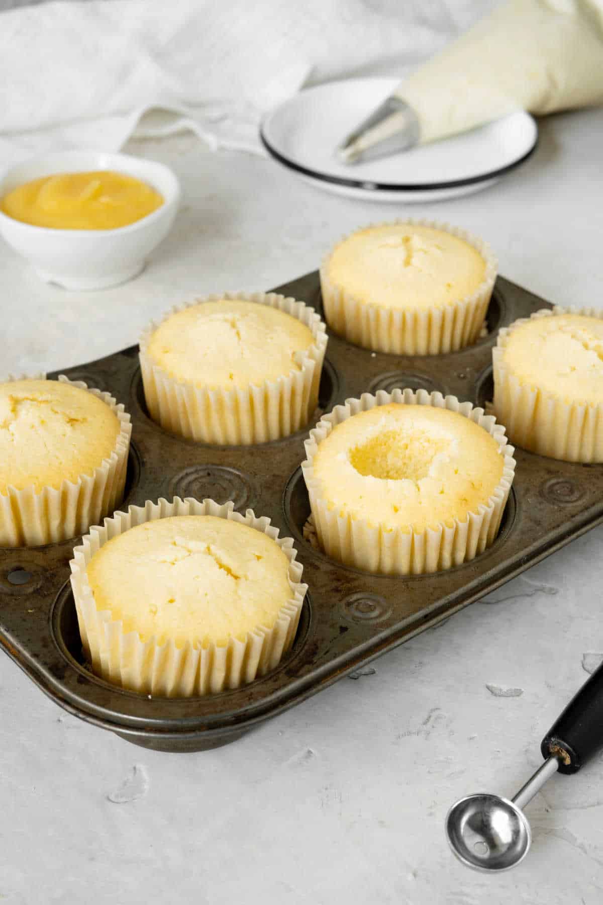 Metal muffin pan with lemon cupcakes, a melon baller, bowl with lemon curd, piping bag. Grey and white surface.