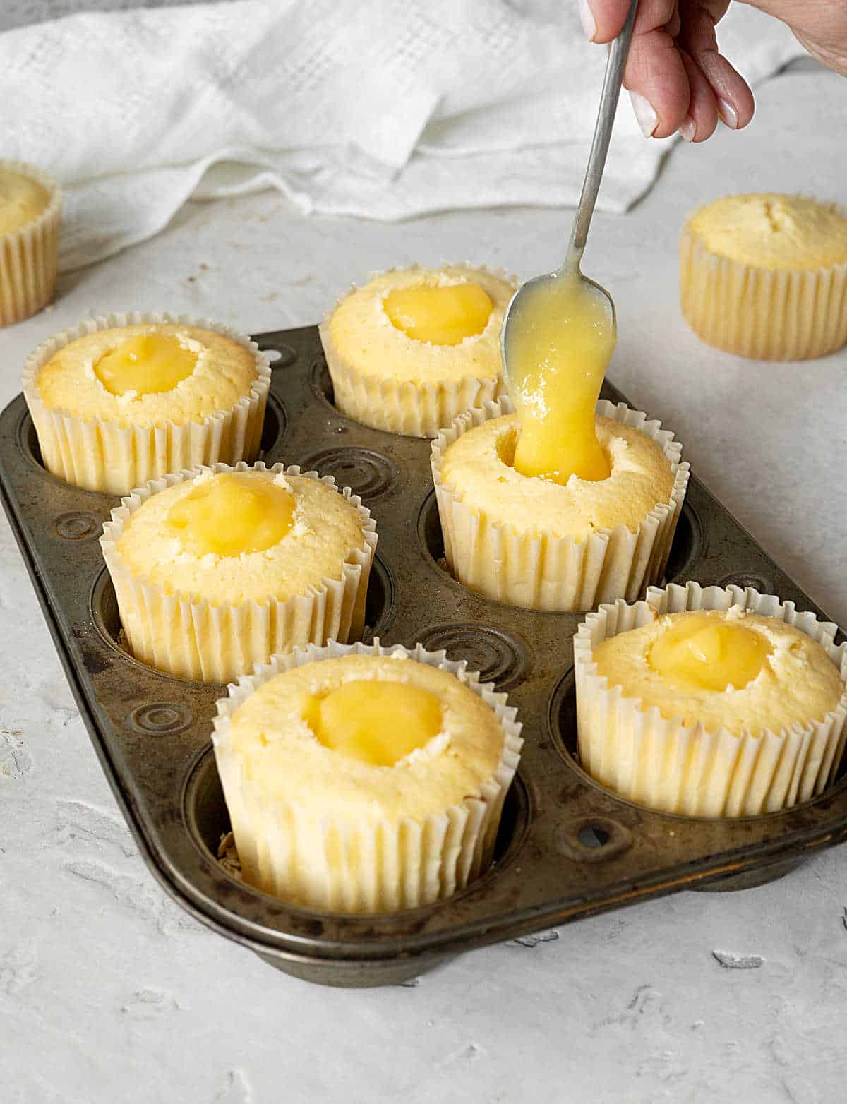 Spooning lemon curd in the center of lemon cupcakes in liners on a metal tin. Grey surface.