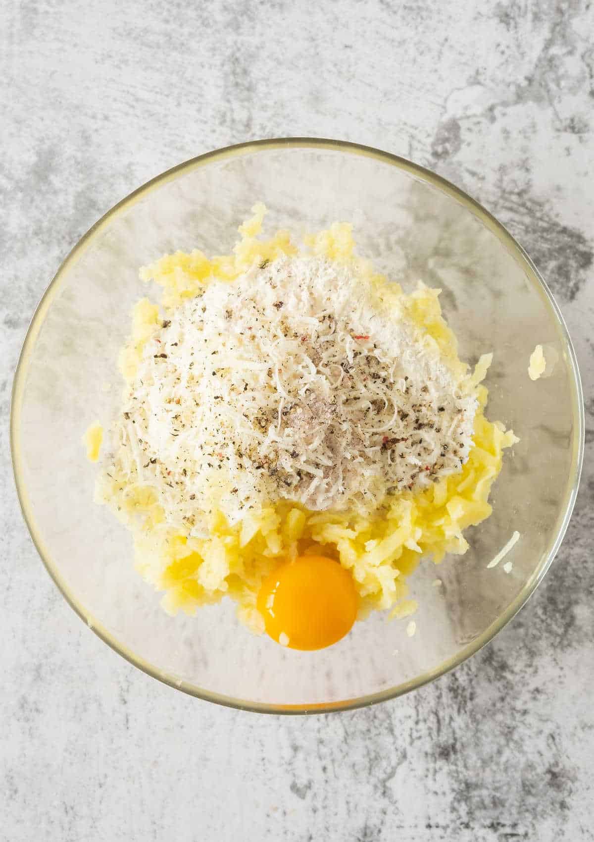 Glass bowl with mashed potato, egg and flour on a gray surface. 
