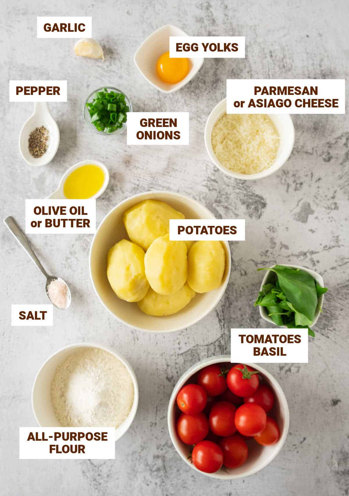 Grey surface with bowls containing ingredients for potato gnocchi including flour, cheese, egg yolk, seasonings, tomatoes and basil.