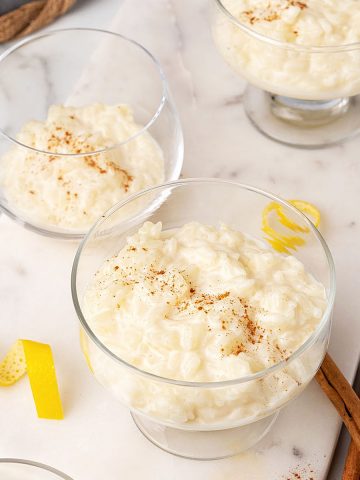 White marble surface with glass bowls containing creamy rice pudding. Cinnamon sticks and lemon peel strips around.