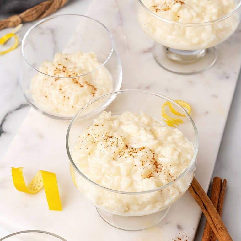 White marble surface with glass bowls containing creamy rice pudding. Cinnamon sticks and lemon peel strips around.