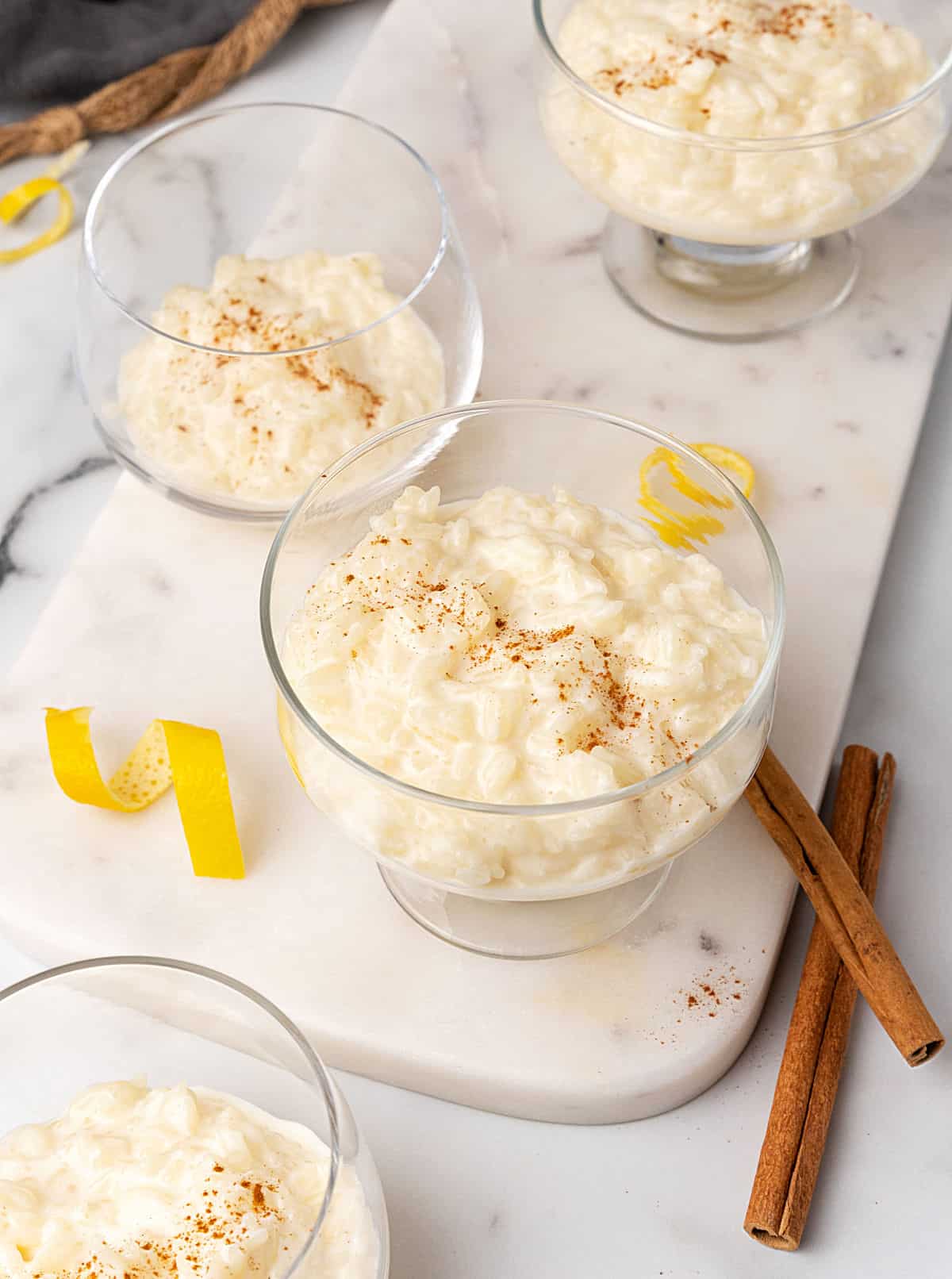 Several rice pudding servings in glass cups on a white marble board. Cinnamon sticks and lemon peel strips around.