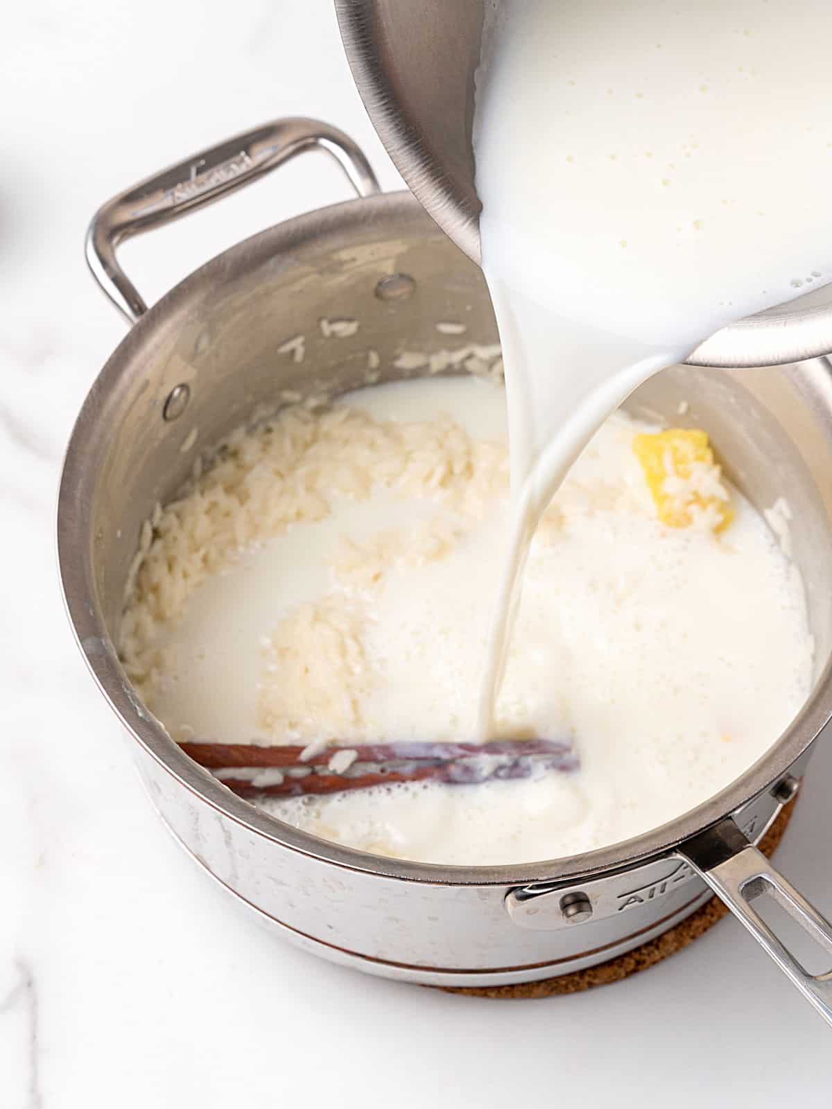 Adding milk to a saucepan with rice, more milk and cinnamon sticks. White marble surface.