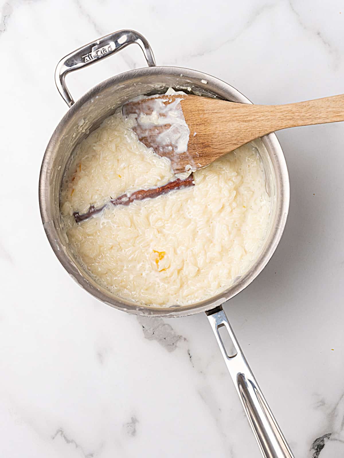 Wooden spoon inside a metal saucepan with cinnamon rice pudding. White marble surface.