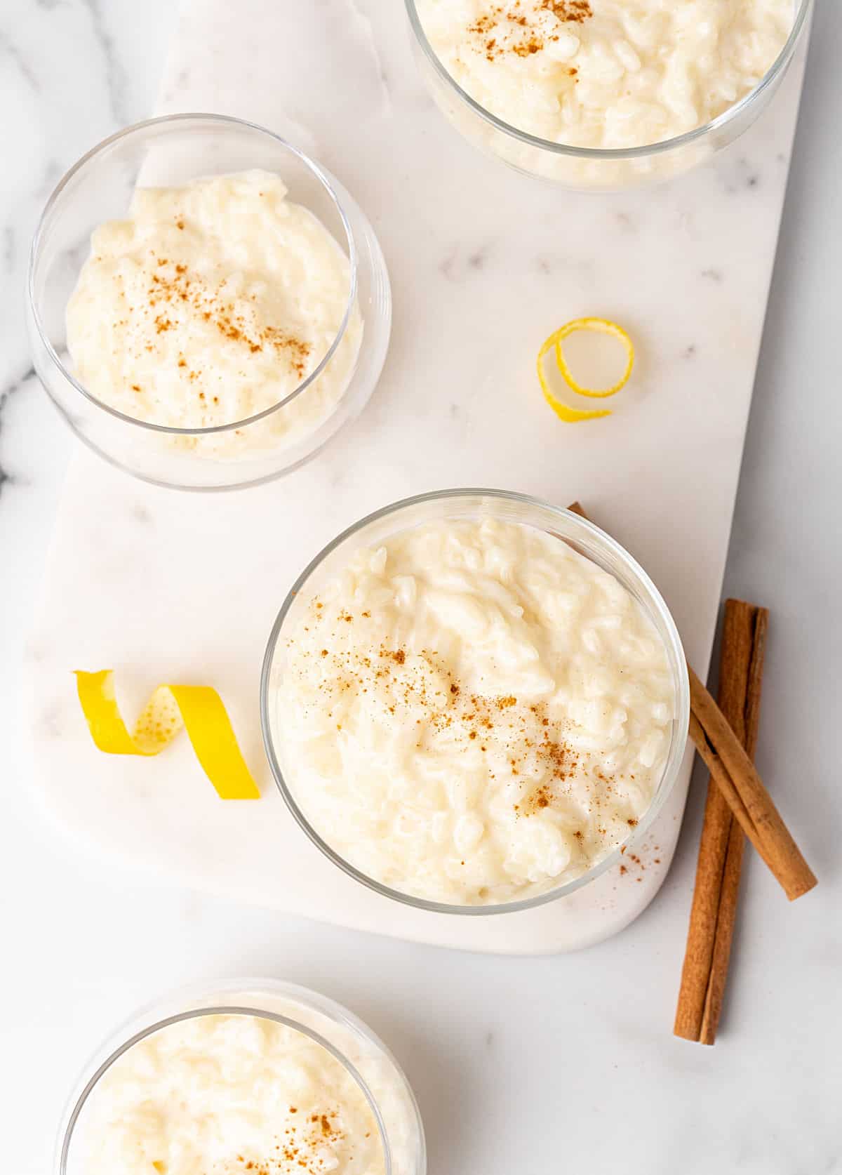 Several glass cups on white marble with rice pudding and cinnamon. Lemon peel strips and cinnamon sticks around. 