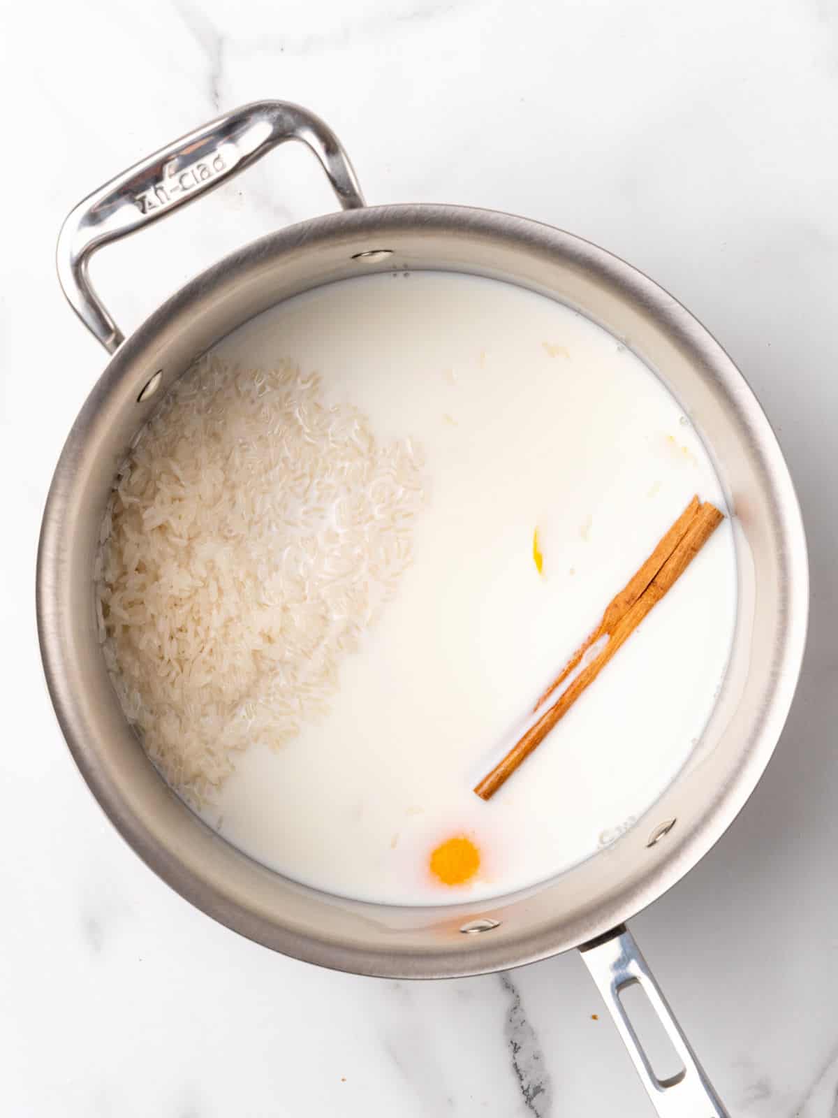 Metal saucepan with milk, rice, cinnamon stick, and citrus peel. White marble surface.