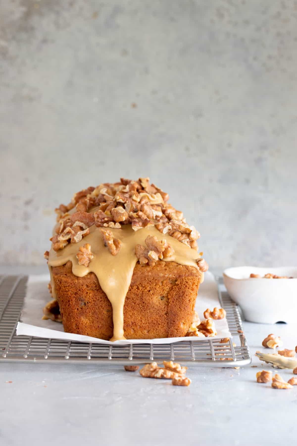 End view of quick bread with glaze and walnuts on top on a wire rack, loose walnut pieces, grey background