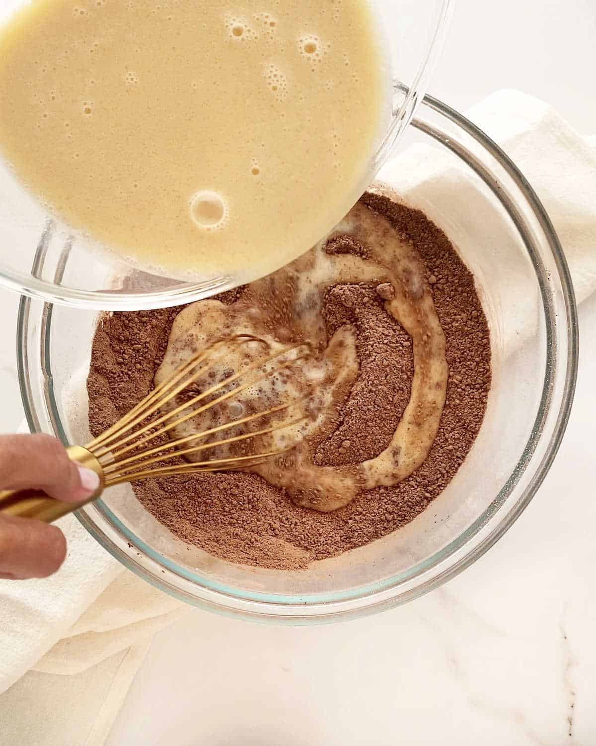 Adding wet ingredients into chocolate cake dry ingredients in a glass bowl on a white surface.