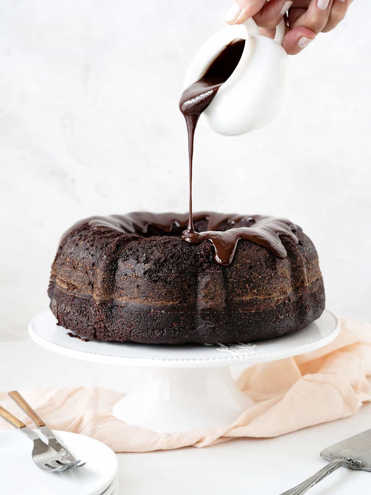 Pouring chocolate ganache from a white sauce boat on a chocolate bundt cake on a white cake stand. Pink cloth underneath, grey background. 