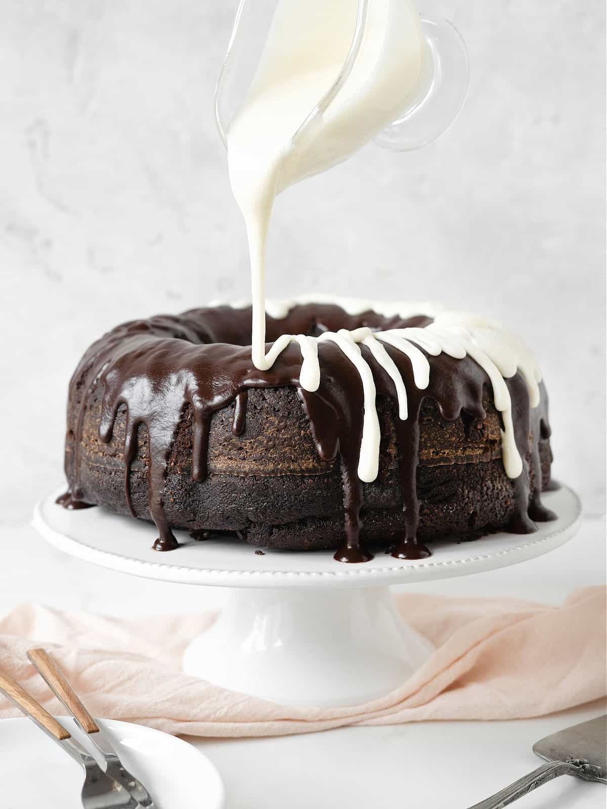 Drizzling cream cheese icing from a pitcher on a chocolate-glazed chocolate bundt cake on a white cake stand. Pink cloth beneath and grey background.