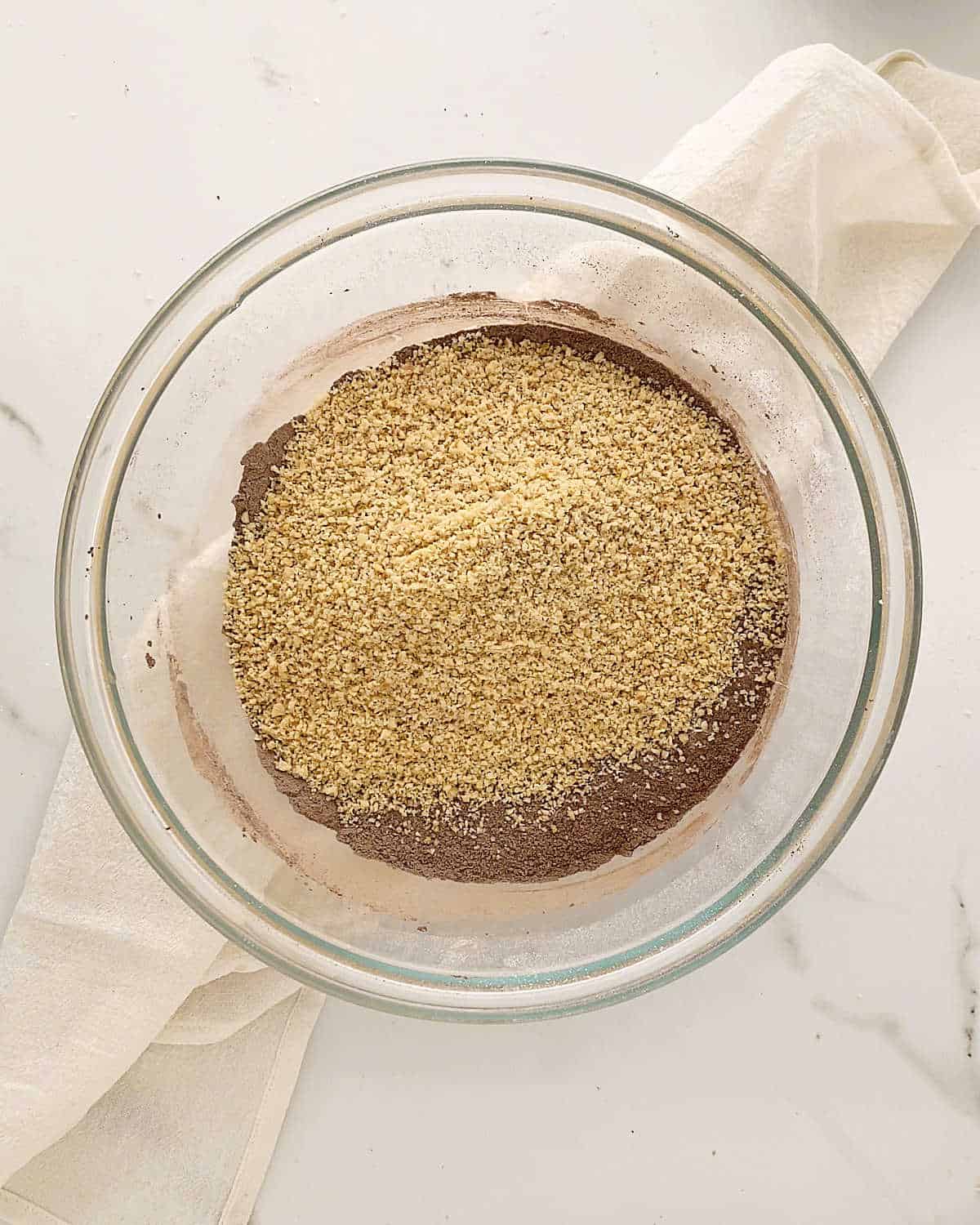 Ground hazelnuts and cocoa powder in a glass bowl set on a white cloth on a white marble surface.
