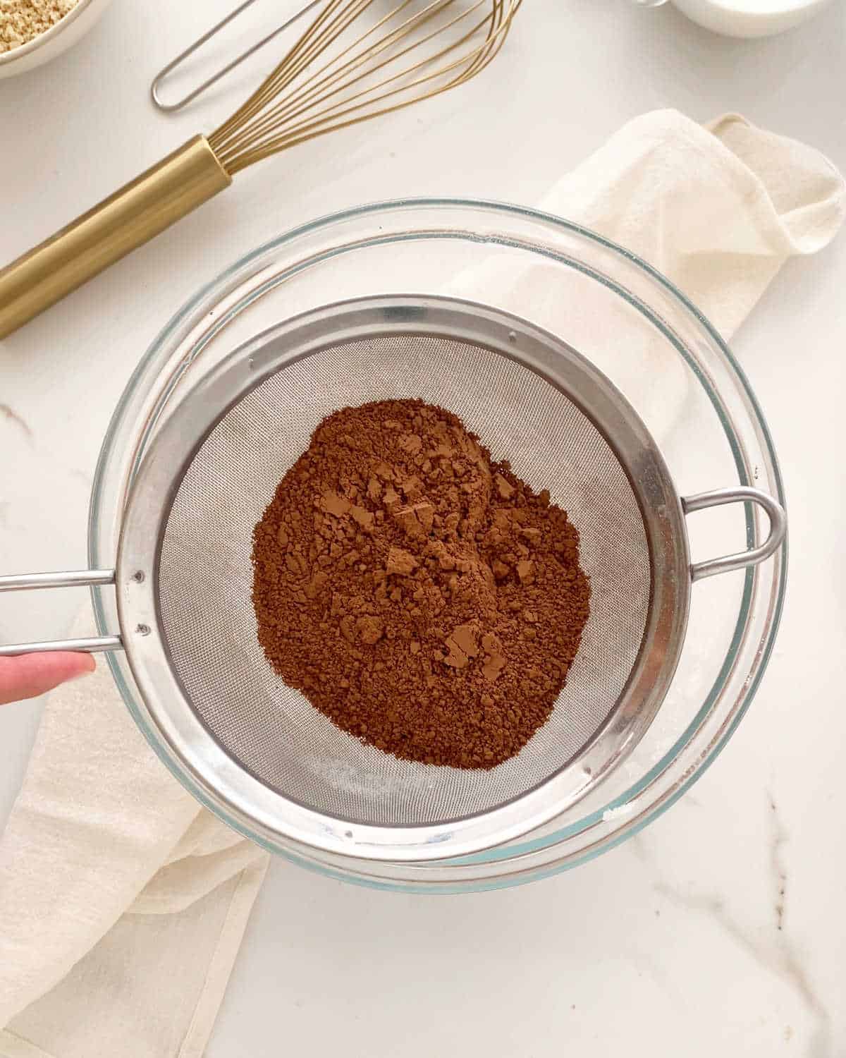Sifting cocoa powder over a glass bowl with a white cloth beneath it on a white marble surface. 