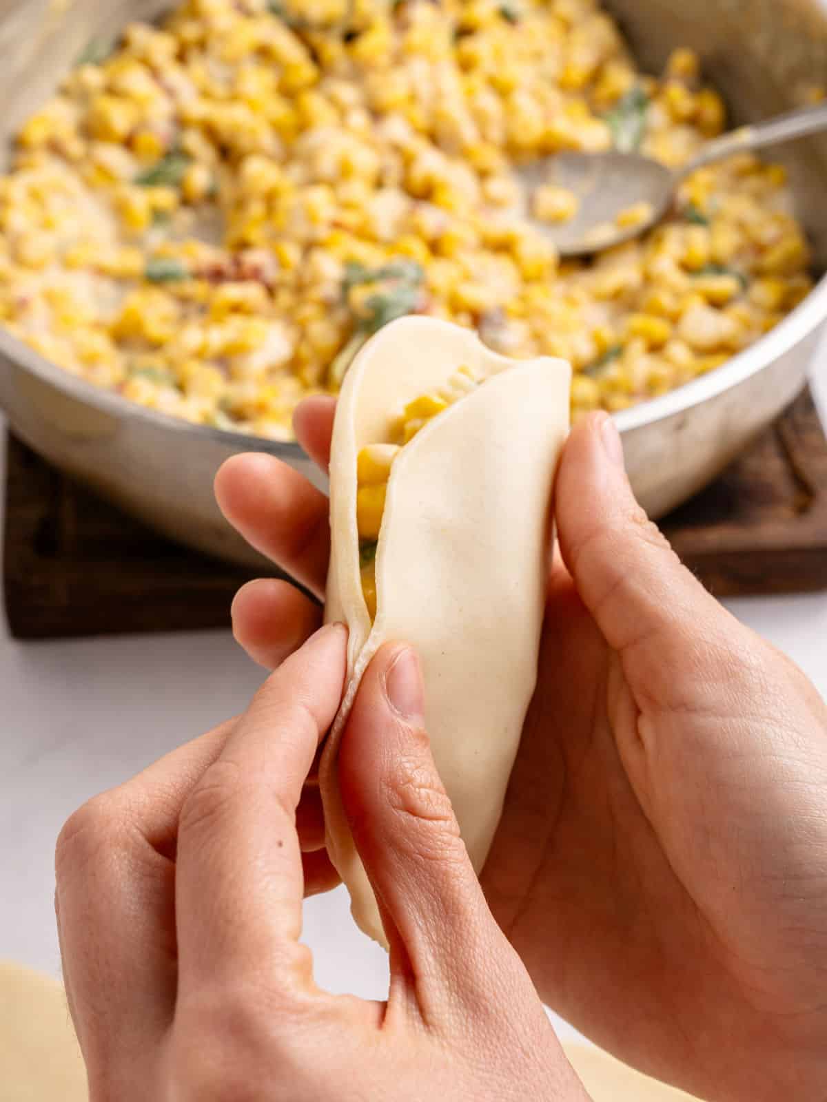 Hands closing corn empanada. Filling in skillet in the background.